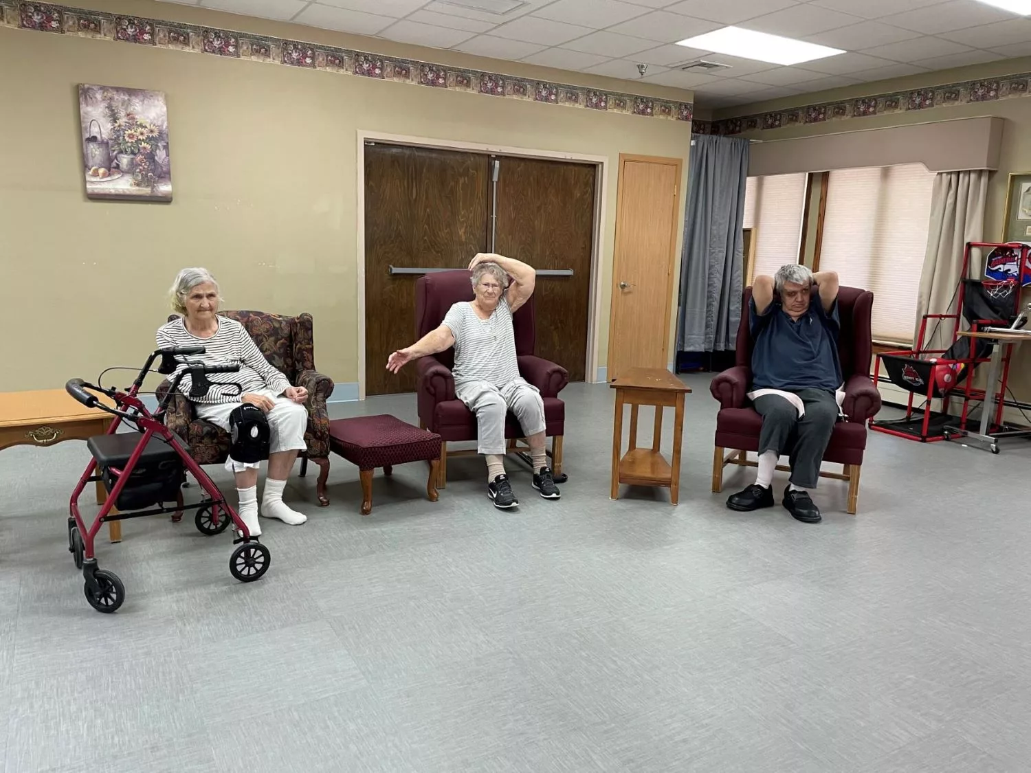 Three seniors exercising indoors with a walker and chairs.