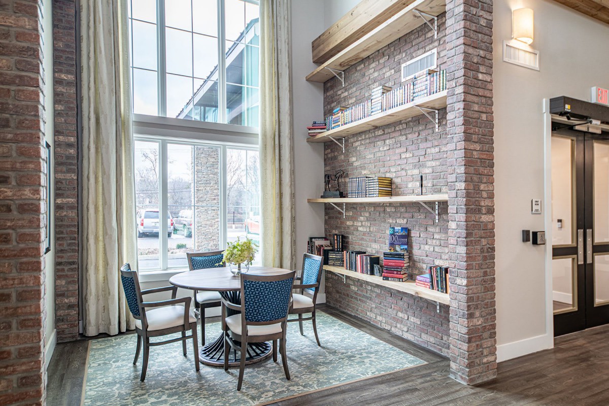 A modern living room with a brick wall, bookshelves, and a cozy table and chairs by large windows.