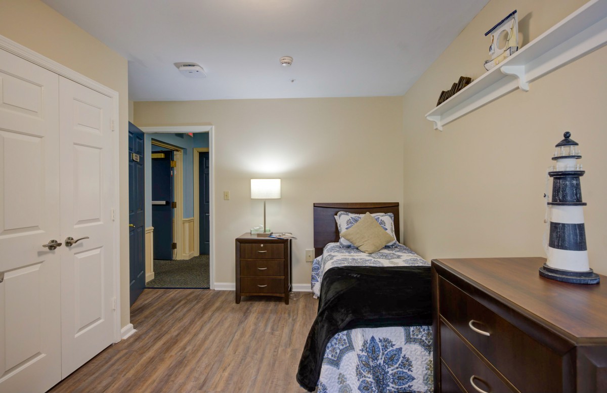 A cozy bedroom with a single bed and wooden furniture, featuring a lamp and simple decor.