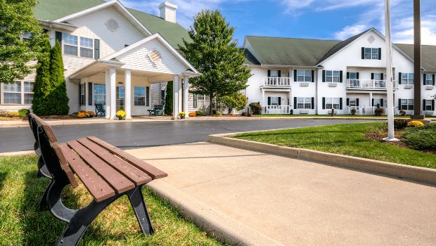 A scenic view of a residential building with a bench and driveway surrounded by greenery.