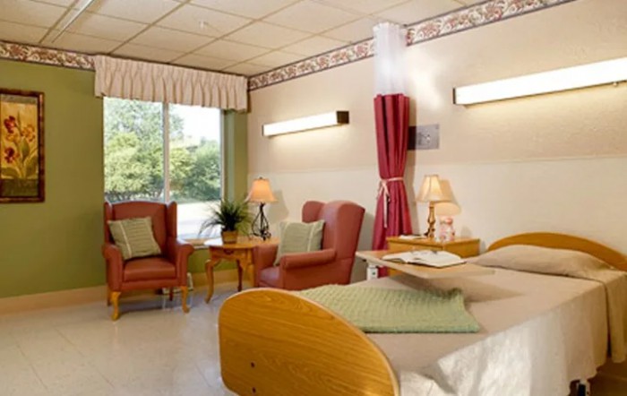 A well-furnished hospital room featuring a bed, seating area, and natural light from the window.