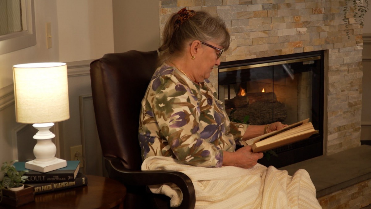 A woman sitting in a chair reading a book by the fireplace.