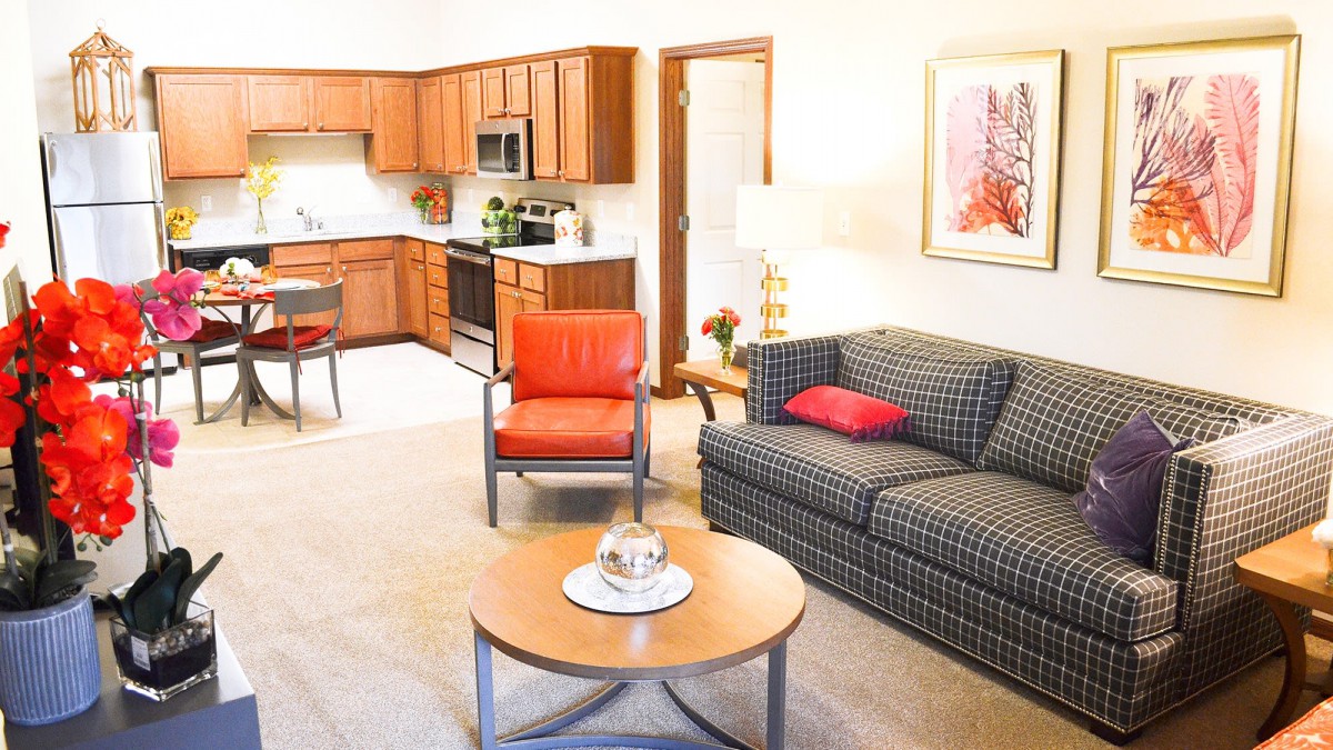 A cozy living room with an open kitchen, featuring a sofa, an orange chair, and a round wooden coffee table.