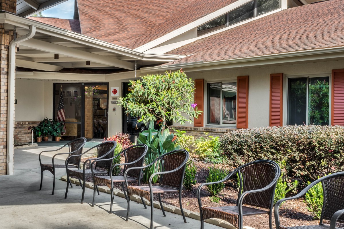 An outdoor seating area with several chairs in front of a building, surrounded by greenery and flowers.