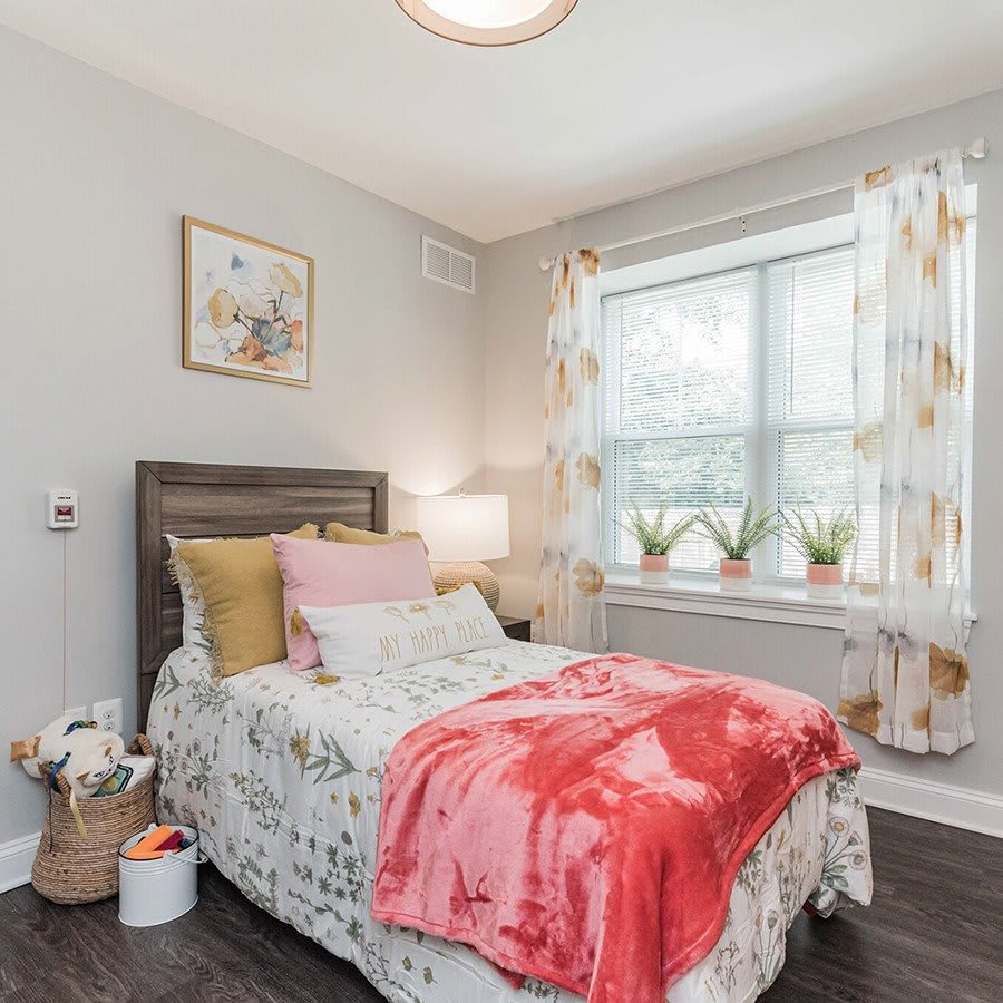 A cozy bedroom with a neatly made bed featuring colorful pillows and a bright blanket, complemented by potted plants on the windowsill.
