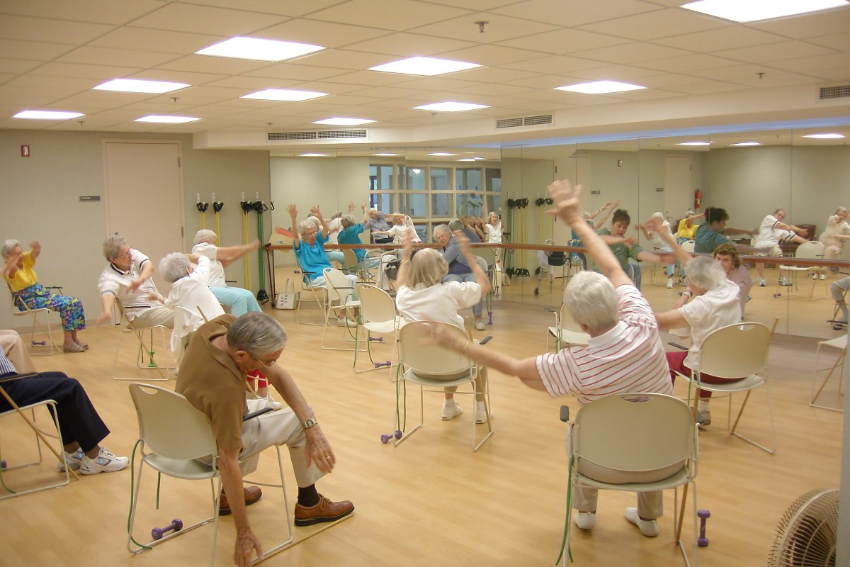 A group of seniors participating in an indoor fitness class where they are performing stretching exercises while seated on chairs.