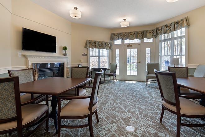 A cozy living room with tables, chairs, a television, and a fireplace near large windows.