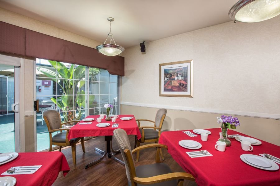 A cozy dining room with tables covered in red tablecloths, set for a meal, featuring comfortable chairs and a bright window view.