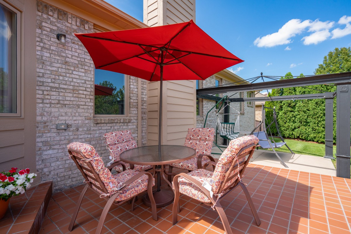A cozy patio setup featuring a red umbrella, round table, and cushioned chairs against a brick wall on a sunny day.