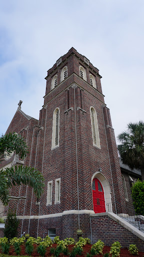 A picturesque brick church featuring a prominent tower and striking red door, surrounded by palm trees.
