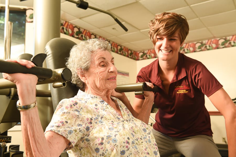 An elderly woman receives assistance from a healthcare worker while using gym equipment.