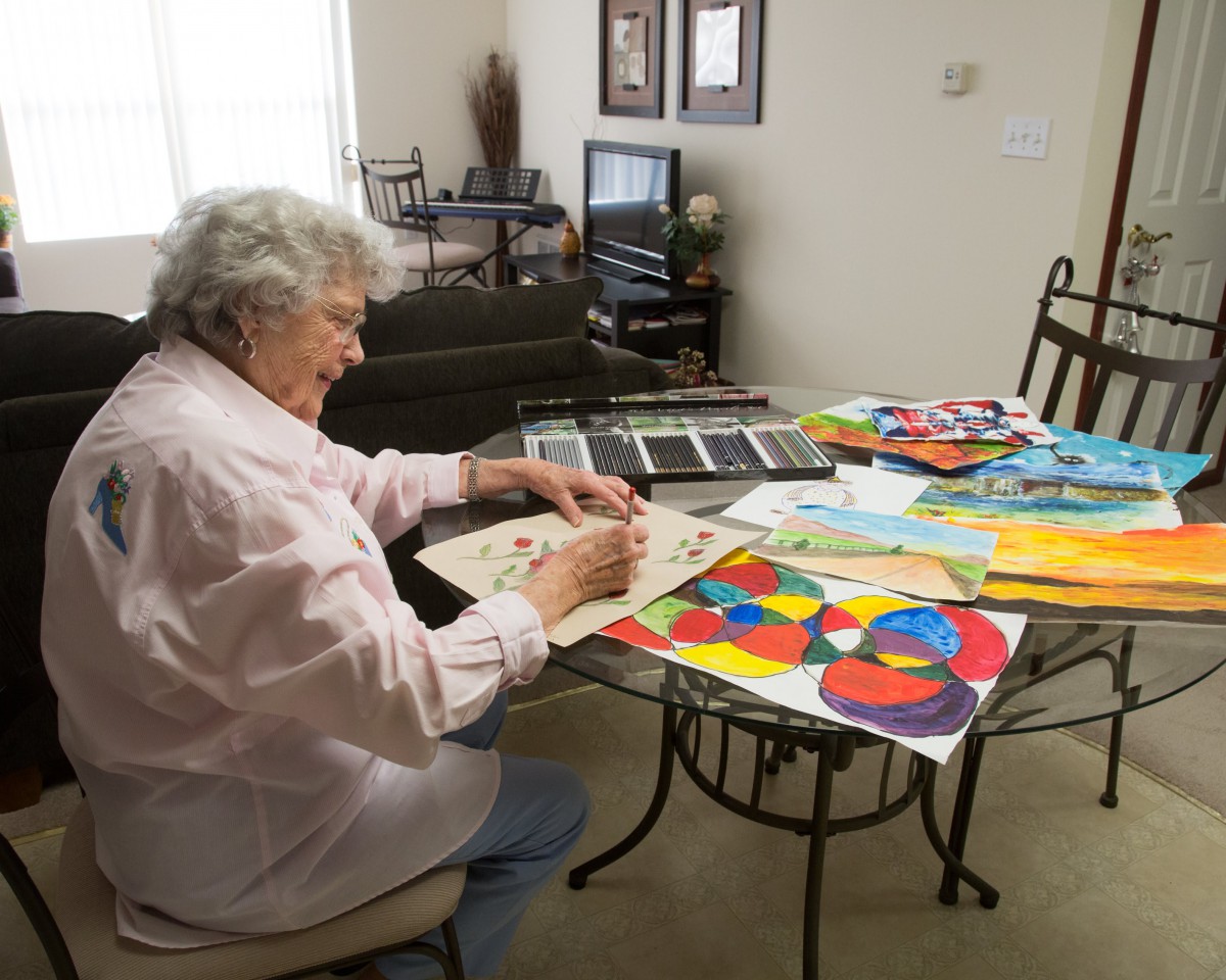 An elderly woman painting colorful artworks at a table indoors.