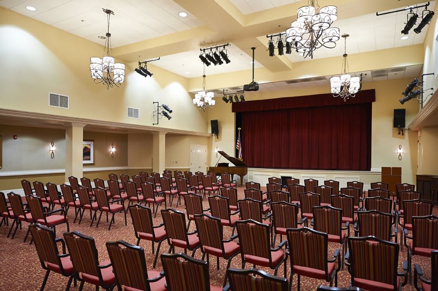 An empty auditorium with rows of chairs, a stage with red curtains, and a piano under elegant chandeliers.