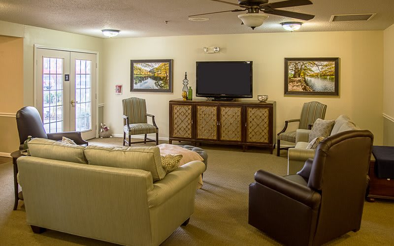 A cozy living room with a sofa, chairs, and a television, decorated with paintings and a ceiling fan.
