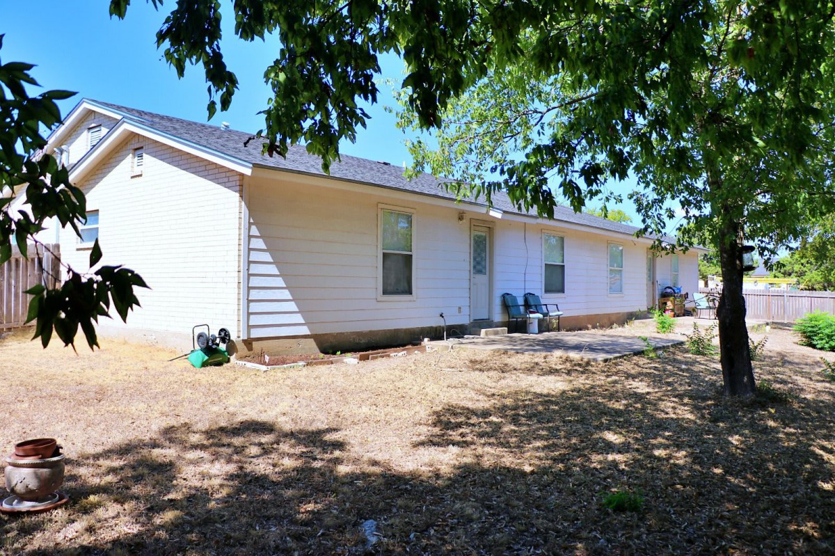 A sunny day view of a one-story house with a yard and chairs under the shade of trees.