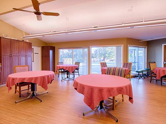 A cozy dining area with tables covered in pink tablecloths, surrounded by large windows and a ceiling fan.