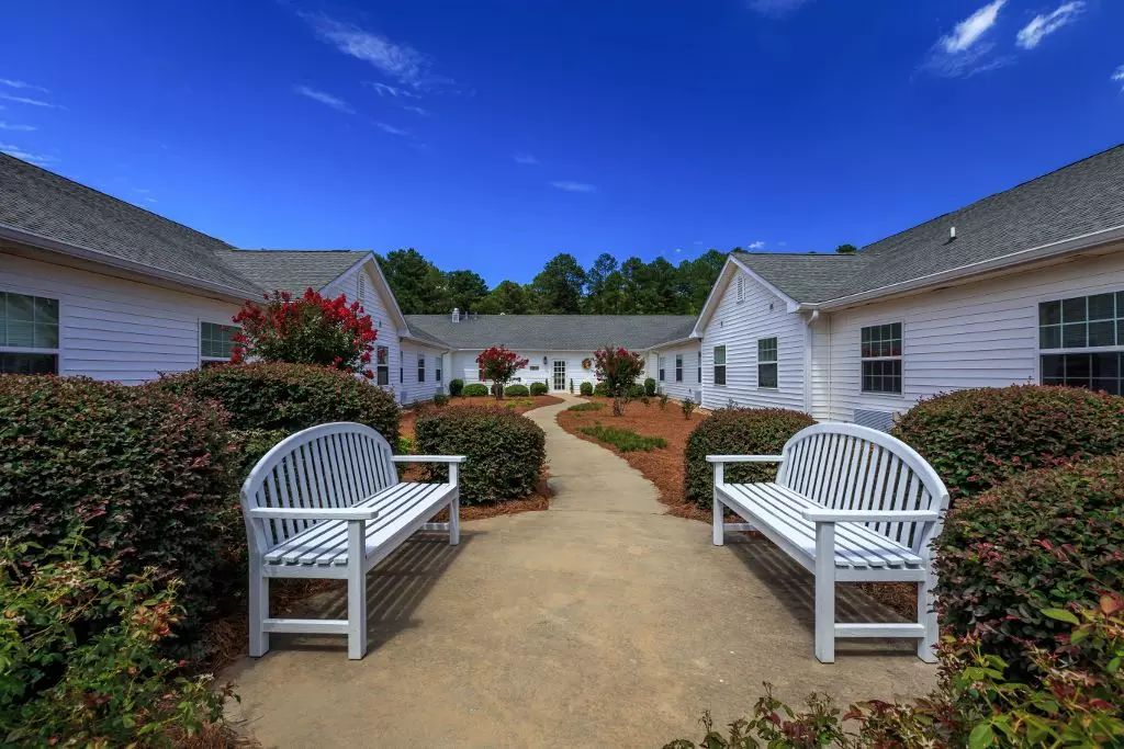 A peaceful courtyard with white benches and well-maintained landscaping, surrounded by white buildings under a clear blue sky.