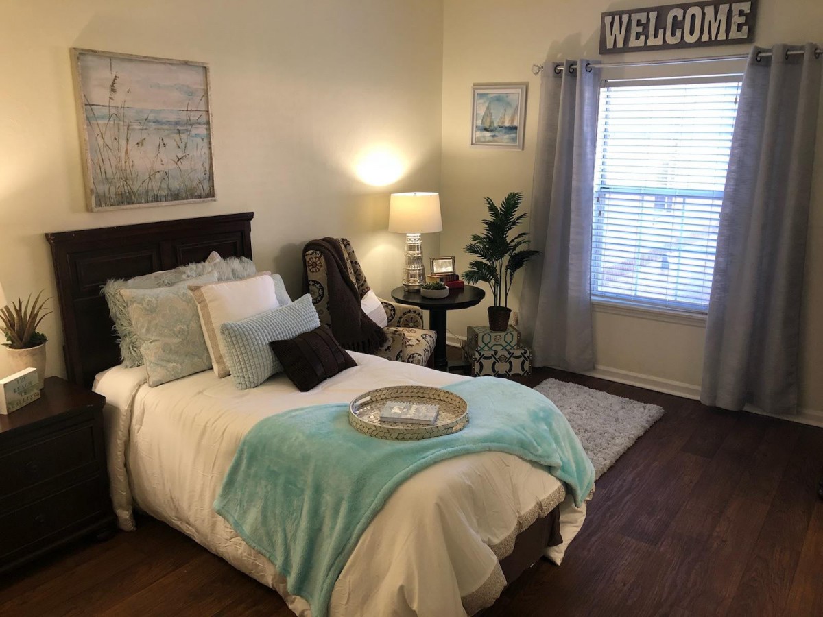 A cozy bedroom with a neatly made bed adorned with soft pillows and a turquoise throw blanket. A bedside table holds a lamp, while a welcome sign hangs above the window.