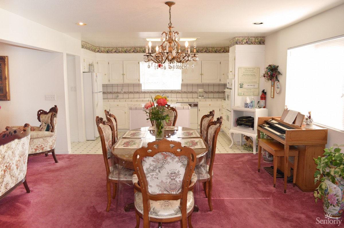 A cozy dining area with a chandelier, round dining table, and chairs on a carpeted floor, adjacent to a kitchen.