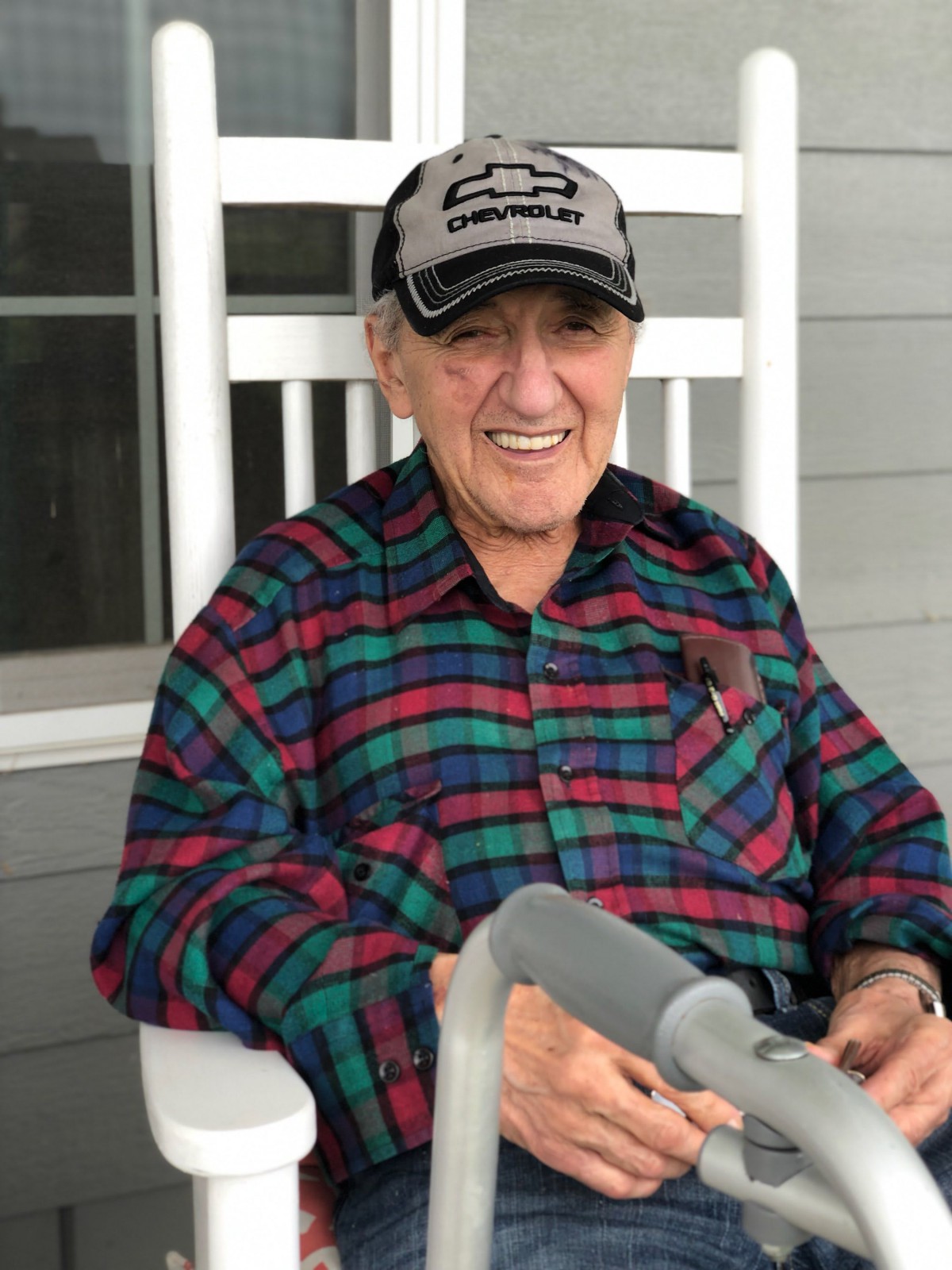 An elderly man wearing a plaid shirt and baseball cap smiles while sitting on a porch.