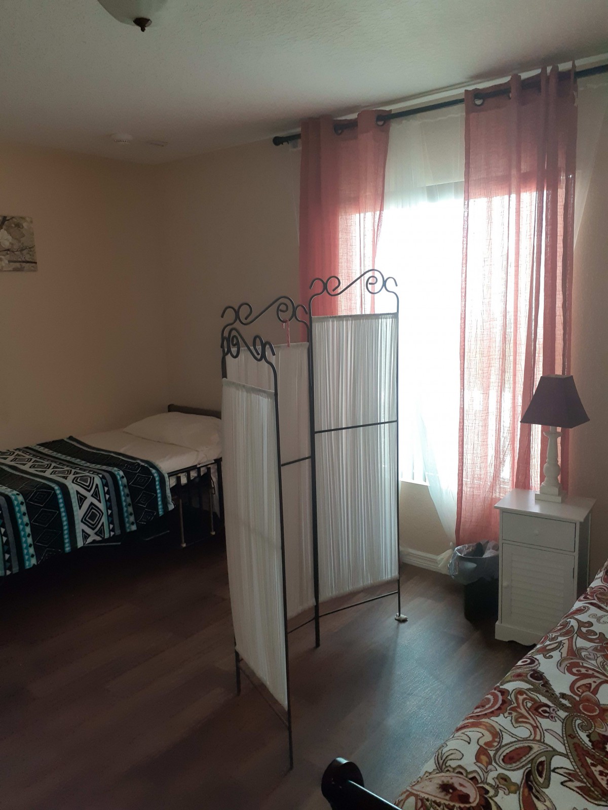 A cozy bedroom featuring two twin beds separated by a decorative room divider, with natural light streaming through pink curtains and a small lamp on a bedside table.