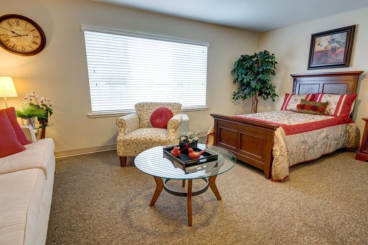 A cozy bedroom featuring an armchair, a glass coffee table, and a bed, with natural light streaming through the window.