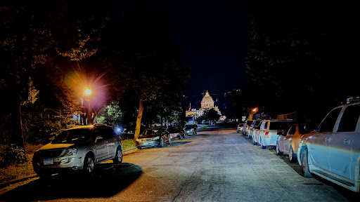 A quiet street scene at night, illuminated by streetlights and lined with parked cars.