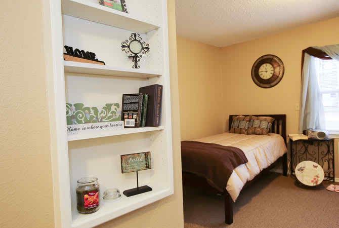 A cozy bedroom featuring a simple bed with brown and beige bedding and a decorative wall shelf with various items.