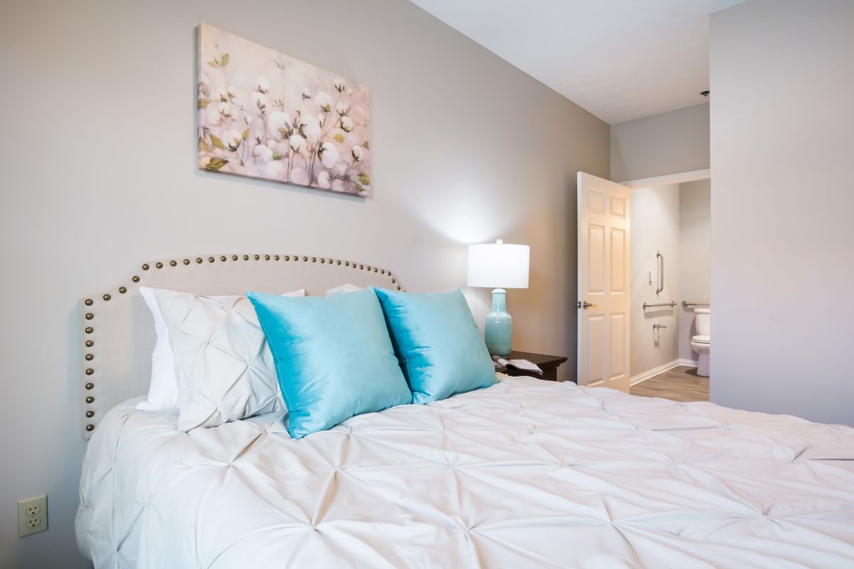A cozy bedroom with a neatly made bed featuring white linens and blue accent pillows, complemented by a bedside lamp and wall art.