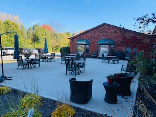 A spacious outdoor patio with tables and chairs, set against a brick building and surrounded by greenery under a clear sky.
