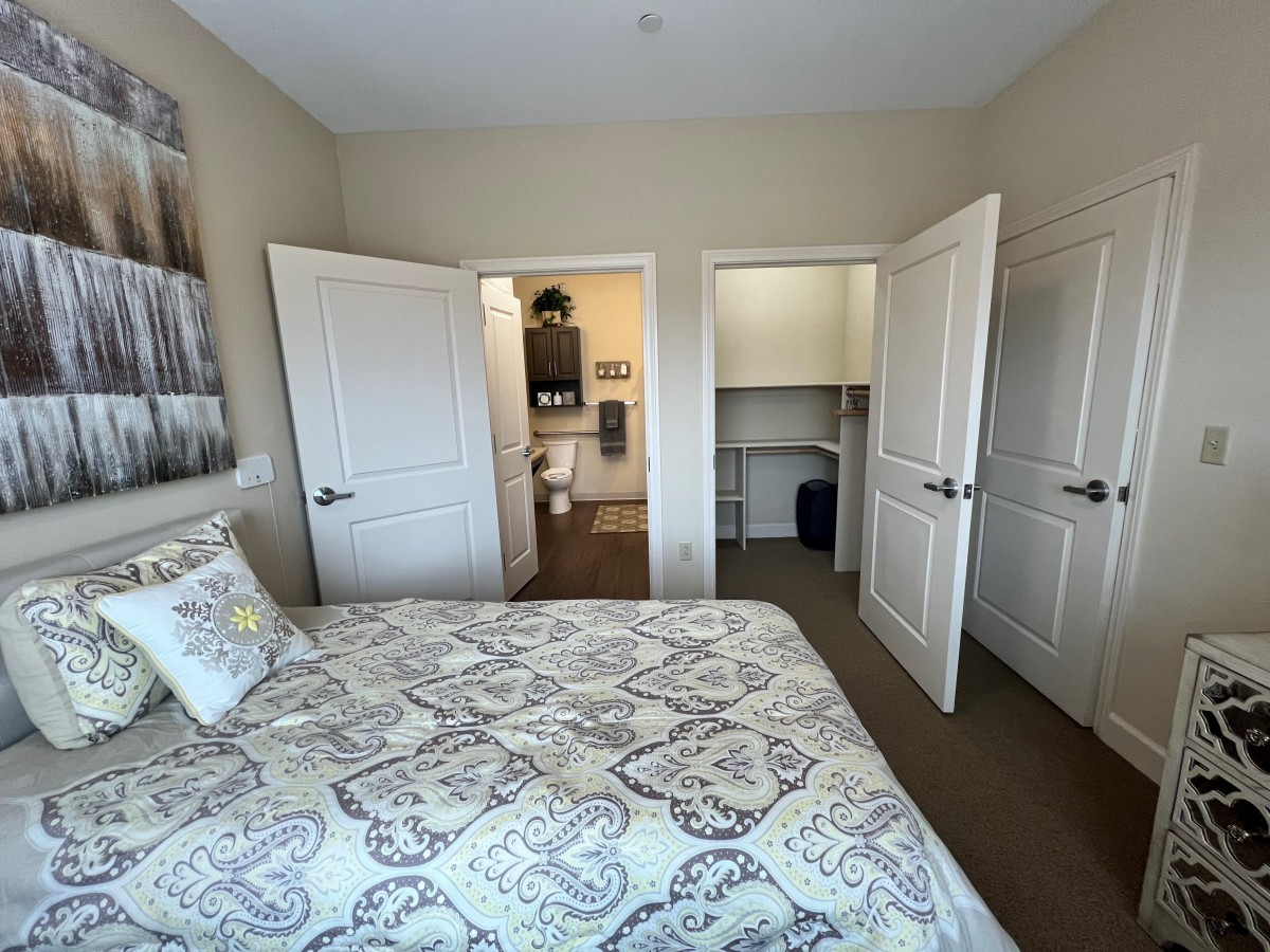 A cozy bedroom with a patterned bedspread, open closet doors, and a glimpse into an adjacent bathroom.