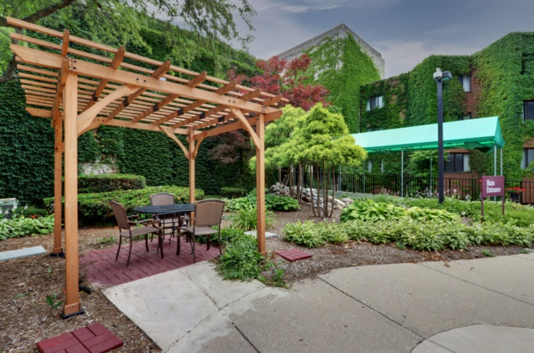 A tranquil garden scene with a pergola and outdoor seating area surrounded by lush greenery and an ivy-covered building.