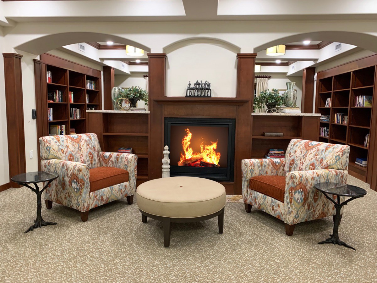 A cozy living room with two patterned chairs facing a warm fireplace, surrounded by bookshelves.