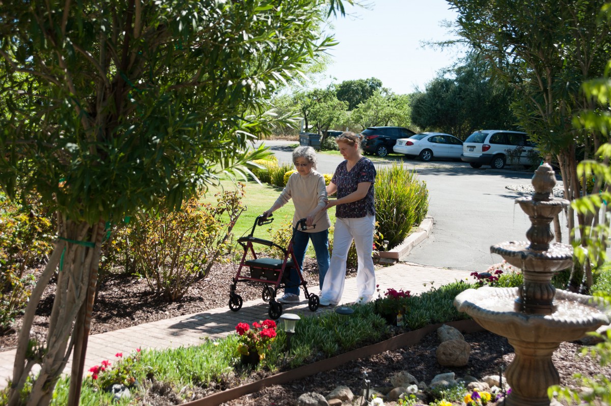 A caregiver assisting an elderly person with walking in a garden setting.