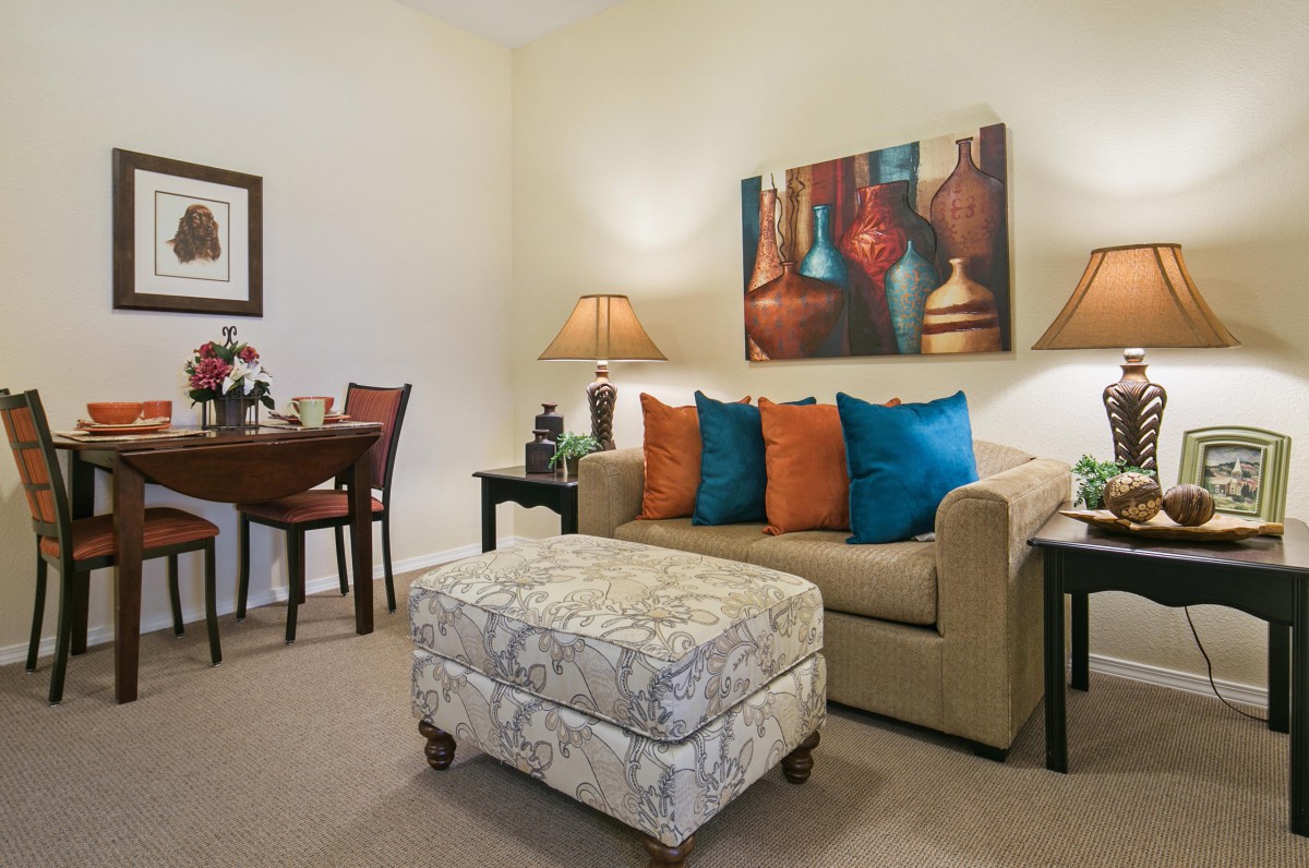 A cozy living room with a beige sofa adorned with colorful pillows, complemented by an ottoman. The space is decorated with modern artwork and ambient lighting from table lamps.
