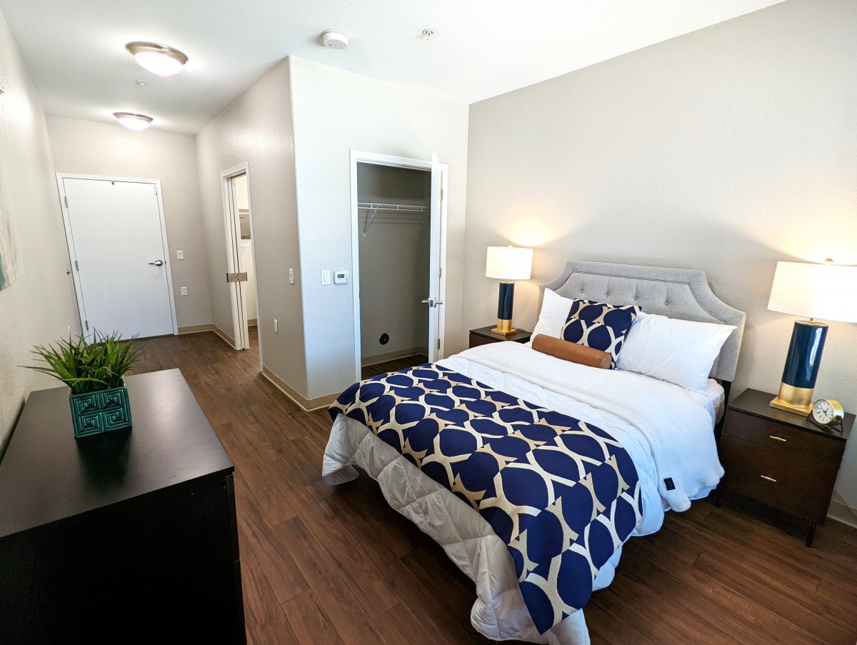 A modern, well-lit bedroom featuring a neatly made bed with patterned bedding, a nightstand with a lamp, and an open closet.