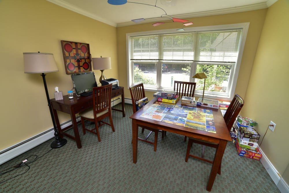 A cozy home office with a desk, chair, and natural light streaming through a large window. A puzzle is laid out on the table.