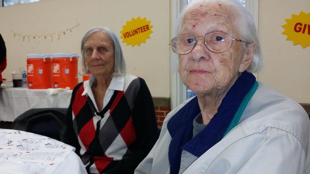 Two elderly individuals are seated indoors, with a 'Volunteer' sign visible in the background.