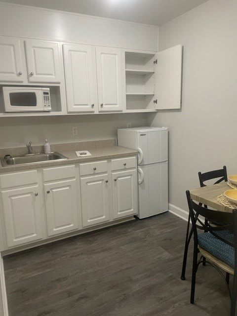 A small kitchen area with white cabinets, a microwave, refrigerator, and a dining table with chairs.