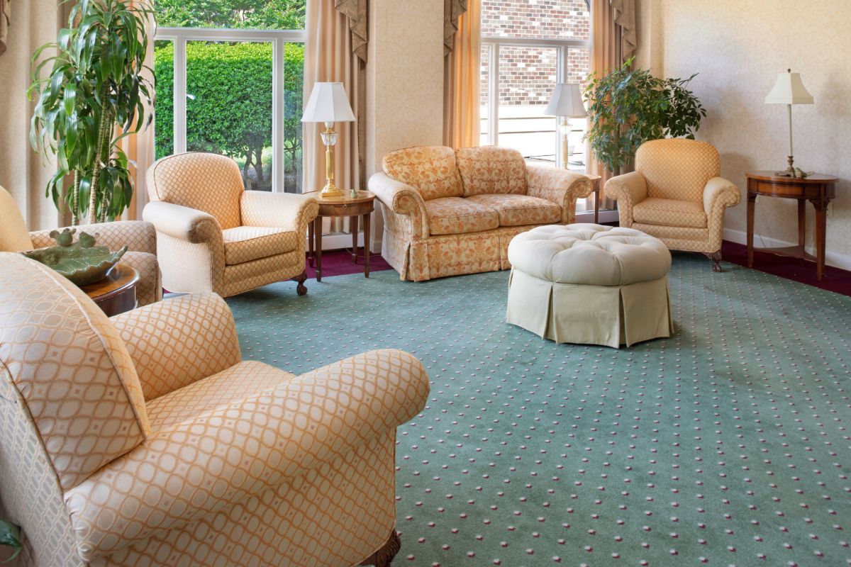 A cozy living room with comfortable sofas and armchairs arranged around a carpeted area, featuring large windows that allow natural light to flood the space.