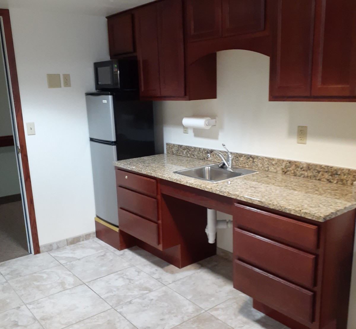 A small kitchen area with wooden cabinets, a compact fridge, and a granite countertop.
