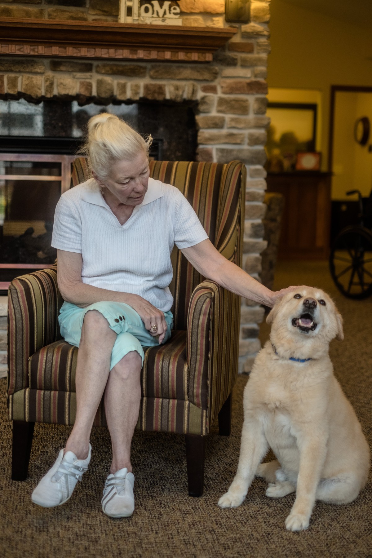 An elderly woman sitting in a chair indoors, smiling and petting a dog.
