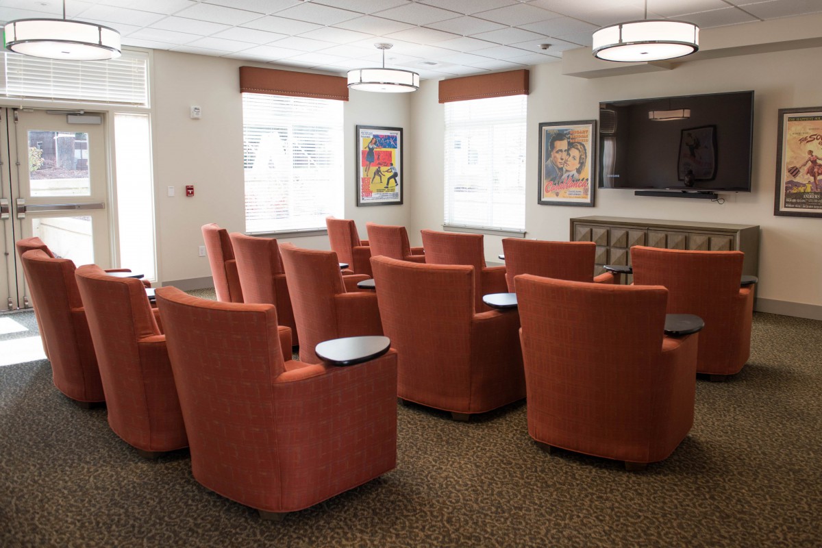 A comfortable seating area with red chairs arranged in rows, featuring a television and large windows for natural light.