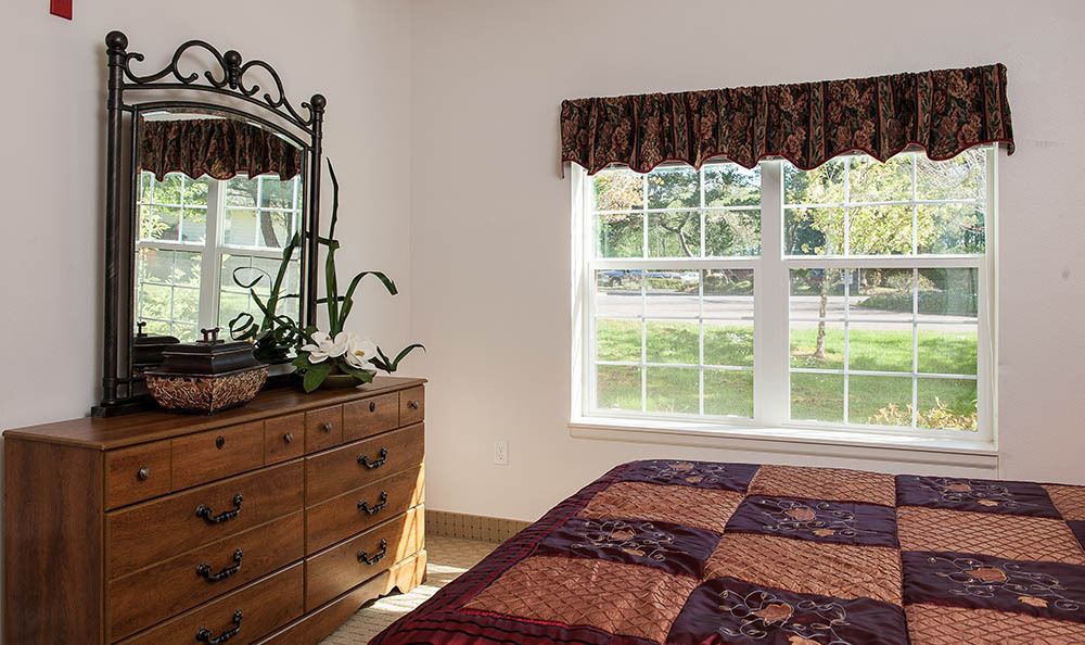 A cozy bedroom with a wooden dresser, mirror, and a bed covered with a colorful quilt next to a sunlit window.