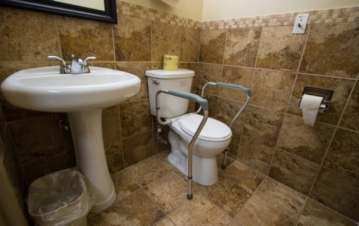 A bathroom with a toilet equipped with handrails for accessibility and a sink, featuring tiled walls and floors.