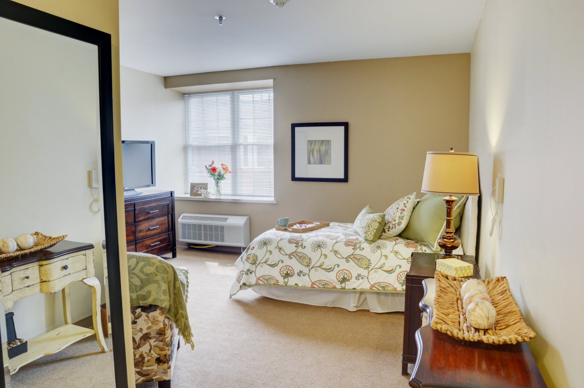 A cozy bedroom with a neatly made bed, bedside lamp, and natural light coming through the window.