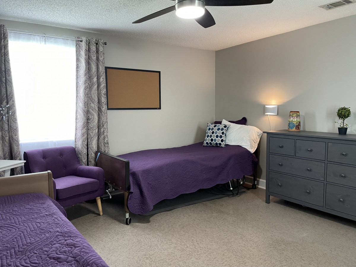 A cozy bedroom with twin beds and purple bedding, featuring a nightstand, dresser, armchair, and ceiling fan.