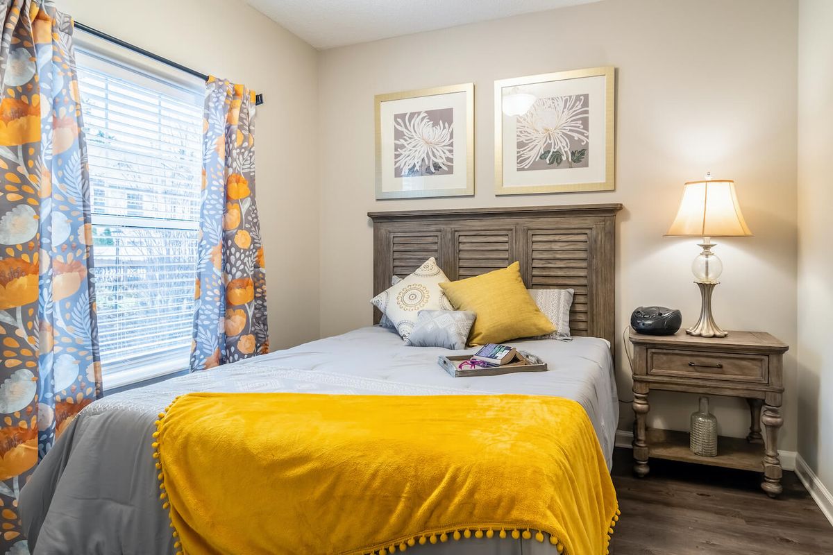 A cozy bedroom featuring a bed with a yellow blanket and decorative pillows, complemented by artwork on the wall and a lamp on the nightstand.
