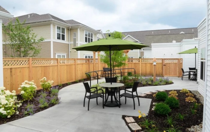 A cozy patio setup with a round table, chairs, and a green umbrella, surrounded by landscaped garden and wooden fencing.