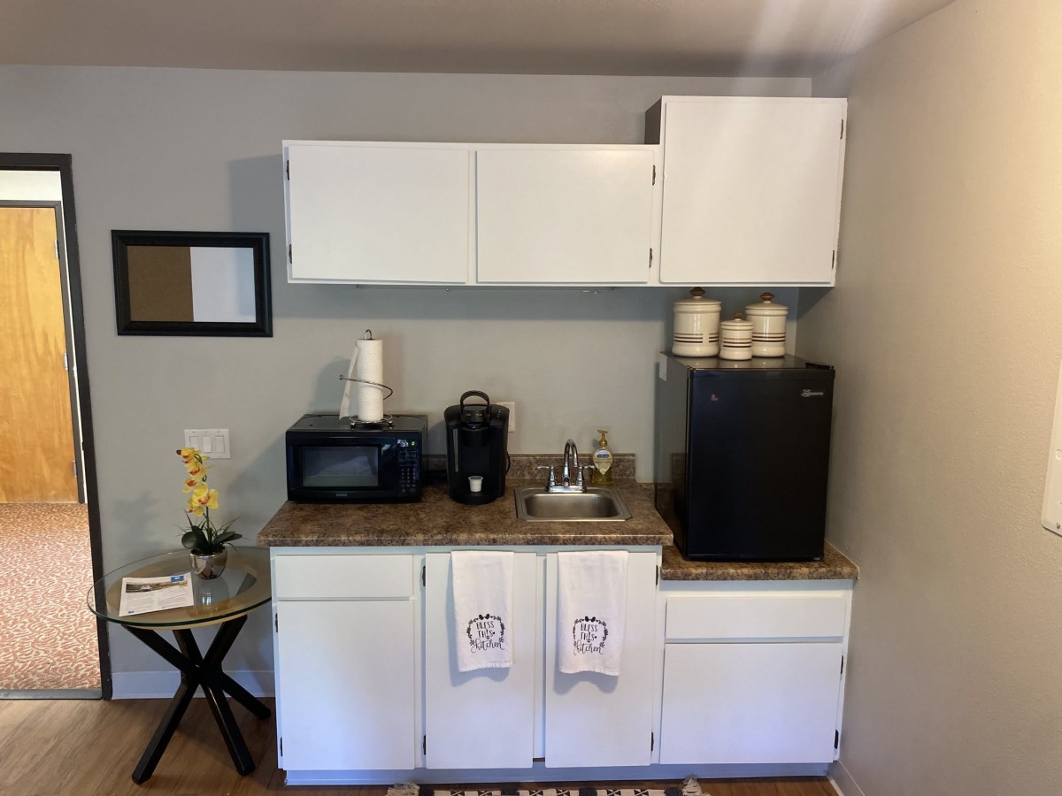 A compact kitchenette with a microwave, coffee maker, mini fridge, and sink, featuring white cabinets and decor.